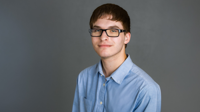 East Texas A&M cybersecurity alum Tag Kalat smiles at the viewer, wearing black-rimmed glasses and a light blue button-up against a gray background.